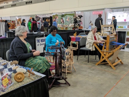 Three people demonstrating spinning wheels and a loom at a craft exhibition.