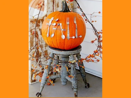 An orange pumpkin decorated with white utensils spelling “FALL,” displayed on a wooden stool.