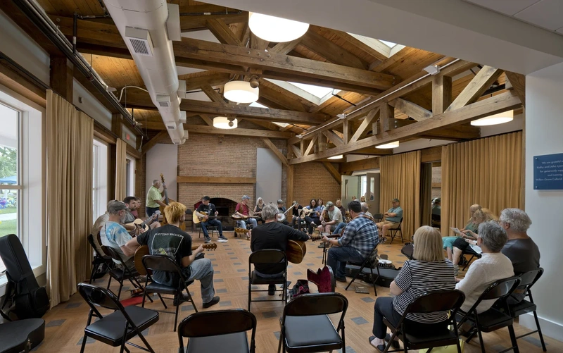 People seated in a circle playing acoustic instruments in a room with exposed wooden beams.