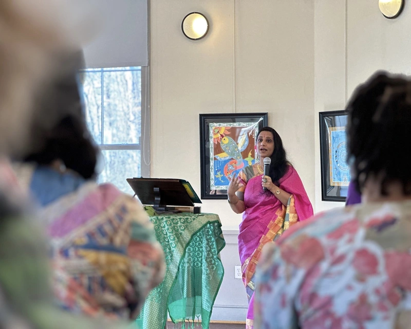 A woman in a pink sari speaks into a microphone at an indoor event, with framed artwork behind her.