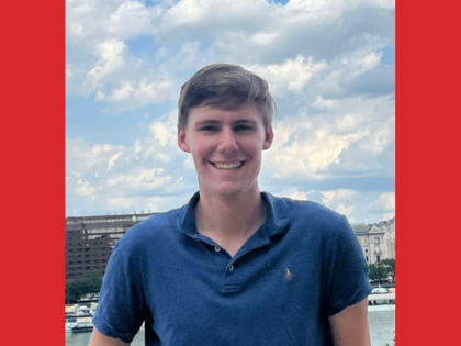 A smiling person outdoors with water and buildings in the background.