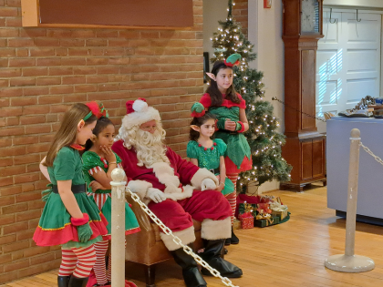 Children in green elf costumes posing with Santa beside a Christmas tree.