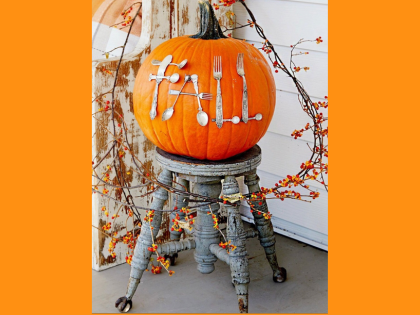 An orange pumpkin decorated with white utensils spelling “FALL,” displayed on a wooden stool.